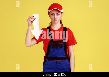 Portrait d'une femme travailleur triste contrariée debout avec une flèche blanche dans les mains indiquant vers le bas, regardant l'appareil photo, portant une combinaison et une casquette rouge. Studio d'intérieur isolé sur fond jaune. Banque D'Images