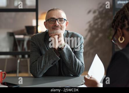 Portrait d'un homme d'affaires mature en lunettes regardant l'appareil photo avec satisfaction tout en étant assis à une table au bureau Banque D'Images