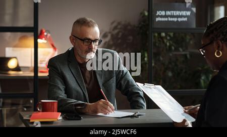 Un gestionnaire sérieux et mature de lunettes assis à la table et examinant le curriculum vitae du candidat pendant l'entrevue d'emploi Banque D'Images