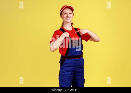 Portrait d'une femme souriante et maniable se tenant debout et se pointant vers elle-même avec une fière expression faciale, se vantant de son succès, portant une combinaison et une casquette rouge. Studio d'intérieur isolé sur fond jaune. Banque D'Images