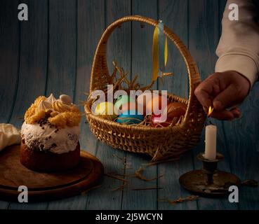 Composition de Pâques. Gâteau de Pâques, un panier avec des œufs de couleur, un porte-bougie en bronze, une main éclairant une bougie avec un fond flou Banque D'Images