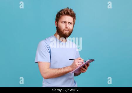 Portrait d'un homme barbu prenant des notes dans un carnet papier, écrivant une idée d'entreprise, des projets d'avenir, regardant loin avec une expression réfléchie. Studio d'intérieur isolé sur fond bleu. Banque D'Images