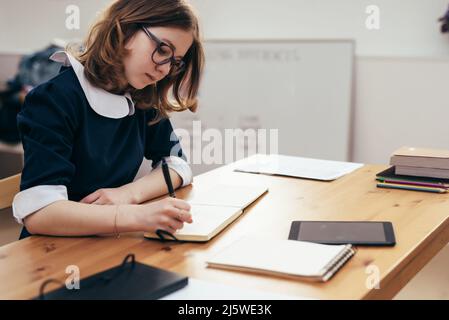 Fille d'école écrit dans un carnet assis à table classwork, devoirs. Banque D'Images