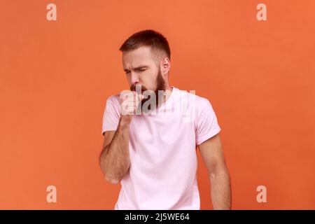 Portrait d'un homme barbu malsain toussant, attrape froid, ayant une température élevée, ayant un symptôme de grippe, portant un T-shirt rose. Studio d'intérieur isolé sur fond orange. Banque D'Images