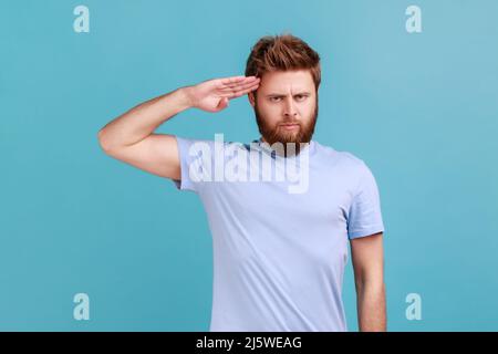 Portrait d'un homme à barbe patriotique responsable saluant avec respect comme si le soldat attendait l'ordre du commandant, obéissant à la discipline. Studio d'intérieur isolé sur fond bleu. Banque D'Images