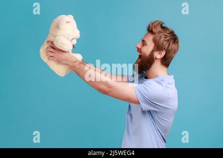 Vue latérale d'un bel homme barbu satisfait debout, tenant un ours en peluche blanc, exprimant des émotions positives, présent romantique. Studio d'intérieur isolé sur fond bleu. Banque D'Images