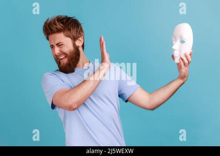 Portrait d'un beau barbu effrayé tenant le masque blanc et le visage tournant, montrant le geste d'arrêt avec la paume, exprimant la peur et le stress. Studio d'intérieur isolé sur fond bleu. Banque D'Images