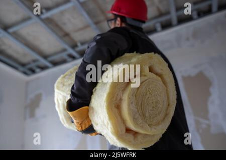 Homme ouvrier en uniforme portant un casque et tenant de la laine de verre pour isoler le grenier sous ses bras. Banque D'Images