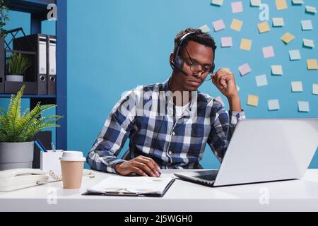 Un télévendeur stressé et fatigué portant un casque tout en essayant d'aider le client de l'entreprise à résoudre son problème. L'opérateur de la ligne d'assistance téléphonique est épuisé et assis à son bureau tout en parlant avec le client. Banque D'Images