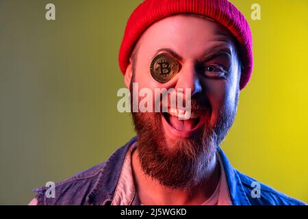 Portrait d'un hipster enjoué et excitant couvrant un œil avec du bitcoin doré, de la crypto-monnaie, portant un bonnet beanie et un gilet en denim.prise en studio en intérieur isolée sur fond coloré de lumière néon. Banque D'Images