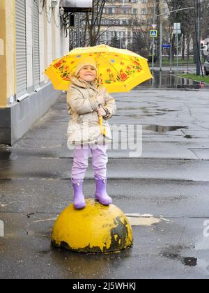 Moscou. Russie. 24 avril 2022. Une fillette heureuse avec un parapluie jaune et des bottes en caoutchouc sur une promenade de printemps le long de la rue de la ville pendant la pluie. Banque D'Images