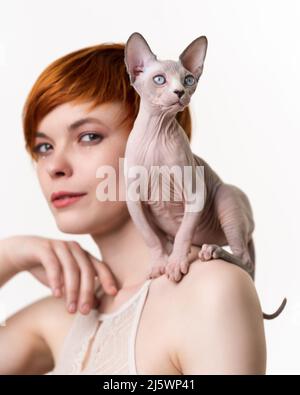 Chat Sphynx canadien assis sur l'épaule d'une jeune femme à tête rouge avec des cheveux courts. Mise au point sélective sur le chat, faible profondeur de champ. Photo de la tête sur blanc Banque D'Images