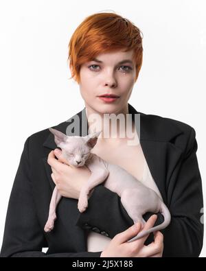 Jeune femme bossy vêtue d'une veste noire tenant les mains dormant chaton Sphynx bleu vison et blanc couleur. Jolie femme à tête rouge avec des cheveux courts Banque D'Images