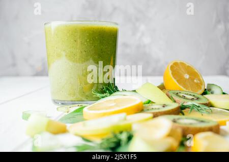 Verre partiellement flou de smoothie vert avec des fruits verts et jaunes et des herbes coupées en morceaux sur la table blanche. Ingrédients pour smoothie. Tranches de Banque D'Images