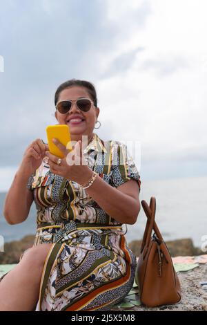 Femme hispanique assise sur le banc vérifiant le téléphone cellulaire dans le parc Banque D'Images