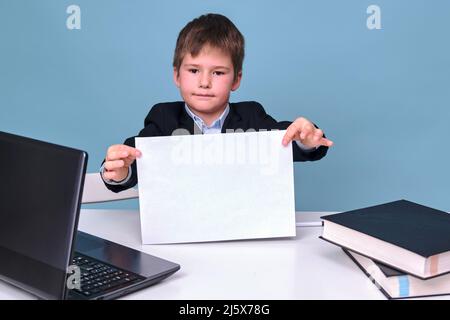 Un garçon dans un costume d'école avec une feuille de papier de A4 à un ordinateur pendant l'apprentissage à distance, un espace de copie sur un fond bleu studio Banque D'Images