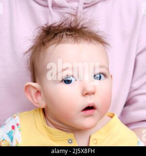 Surpris bébé garçon avec de grands yeux bleus et des cheveux au lave-vaisselle dans ses bras de mère. Portrait d'un enfant effrayé avec une coiffure désordonnée Banque D'Images