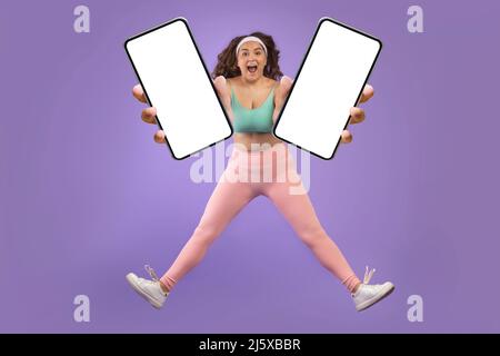 Joyeux jeune caucasien excitée plus de la taille femme dans les vêtements de sport sauter et geler dans l'air, montrer deux smartphones Banque D'Images