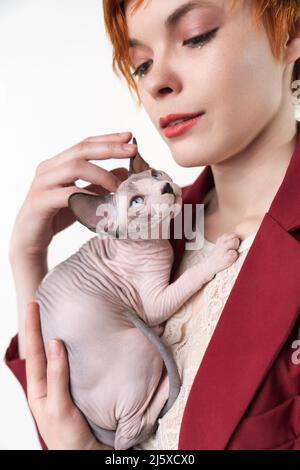 Hipster jeune femme à tête rouge avec des cheveux courts qui embrasse doucement sa poitrine Canadian Sphynx Cat Jolie femme qui regarde loin, vêtue d'une veste rouge Banque D'Images