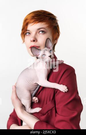 Chat Sphynx canadien assis sur l'épaule de la jeune fille à tête rouge de l'hippopotame. Mise au point sélective sur le chaton domestique sans poils au premier plan, faible profondeur de champ Banque D'Images