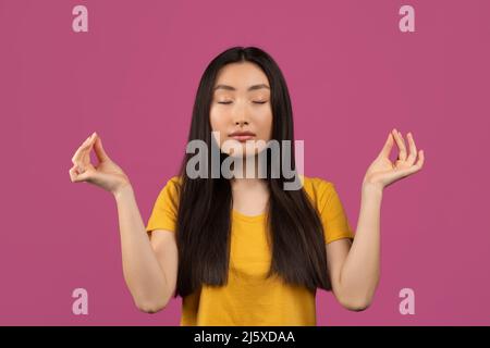 Sereine jeune femme asiatique aux yeux fermés méditant, appréciant le silence et la détente sur fond de studio violet Banque D'Images