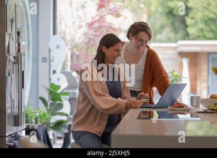 Des amies joyeuses magasinent en ligne sur un ordinateur portable dans la cuisine Banque D'Images