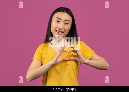 Bonne femme asiatique montrant le geste de coeur avec les mains près de la poitrine, faisant signe d'amour, posant sur fond violet studio Banque D'Images