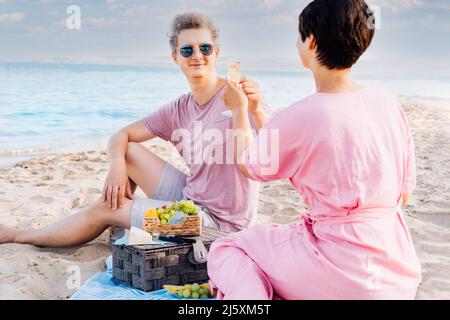 Couple ayant un pique-nique à la plage de bord de mer. Homme et femme amoureux tenant des verres avec du vin ou du champagne, célébrant le moment avec fond de mer. Banque D'Images