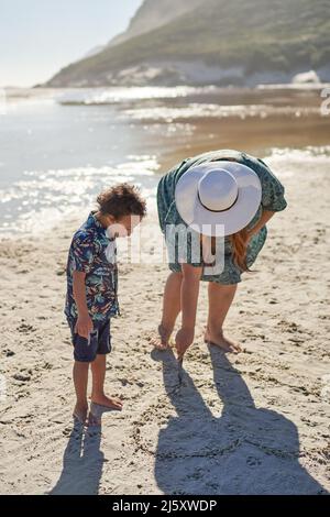 Mère et fils dessinant le coeur dans le sable sur la plage ensoleillée Banque D'Images