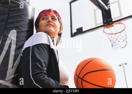 Du dessous de la joueuse avec des cheveux teints dans des vêtements de sport regardant loin avec soin avec le ballon à la main contre le fond de panier de basket-ball Banque D'Images