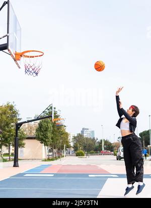 Vue latérale d'une joueuse qui jette le ballon dans un panier tout en jouant au basket-ball contre un ciel nuageux et des arbres verts Banque D'Images