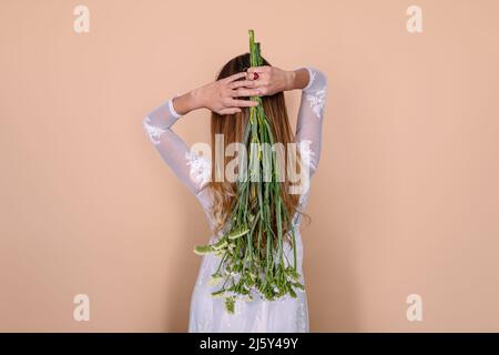 Vue arrière d'une mariée méconnue dans une élégante robe de mariage blanche avec bouquet de fleurs fraîches sur fond beige dans un studio lumineux Banque D'Images