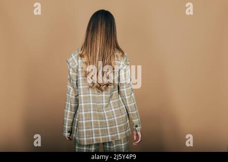 Vue arrière du modèle femelle anonyme avec de longs cheveux ondulés portant une veste à carreaux en studio sur fond beige Banque D'Images