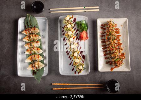 Vue de dessus ensemble de délicieux sushis traditionnels avec sauce et boulettes chinoises servies sur des assiettes blanches près des baguettes et de la sauce soja dans des bols sur bac gris Banque D'Images