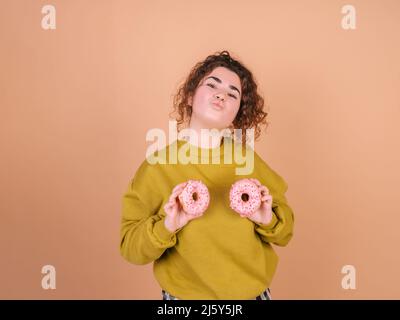 Jeune modèle féminin ludique aux cheveux bouclés faisant un visage de baiser et montrant des beignets doux aux reflets roses en studio et regardant l'appareil photo contre un dos beige Banque D'Images