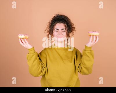 Jeune modèle féminin ludique aux cheveux bouclés montrant des beignets doux avec des reflets roses tout en méditant avec les yeux fermés dans le studio sur fond beige Banque D'Images