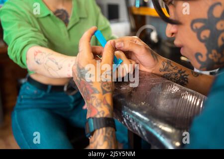 Crop tatoué homme se raser le doigt de la cliente anonyme avec rasoir tout en se préparant à faire le tatouage dans le salon de lumière pendant le rendez-vous Banque D'Images