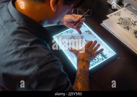 Du dessus de la récolte maître tatoué anonyme faisant tatouage tirage sur tablette tout en étant assis à table dans un salon professionnel de lumière Banque D'Images