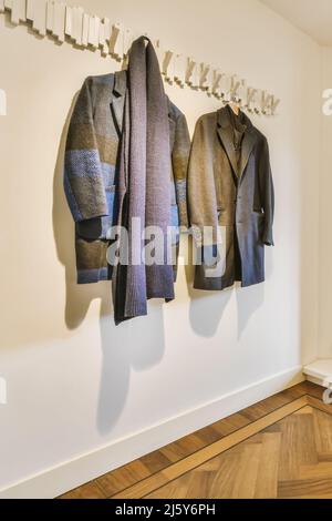 Manteaux gris tendance pour hommes avec foulard foncé élégant suspendu sur des crochets au mur blanc dans la pièce lumineuse de l'appartement Banque D'Images