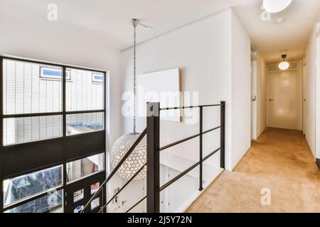 Long couloir avec portes fermées et rambarde noire d'escalier dans un appartement moderne lumineux avec lustre rond et fenêtre Banque D'Images