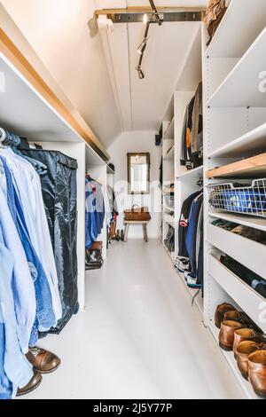 Longue garde-robe légère avec beaucoup de vêtements élégants accrochés sur des étagères blanches contre le miroir dans l'appartement Banque D'Images