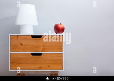 Petite table élégante avec tiroirs et lampe en bois et pomme rouge mûre sur fond gris dans une pièce lumineuse Banque D'Images