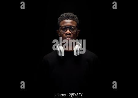 Jeune Afro-américain en lunettes avec casque sur le cou et regardant l'appareil photo sur fond noir Banque D'Images
