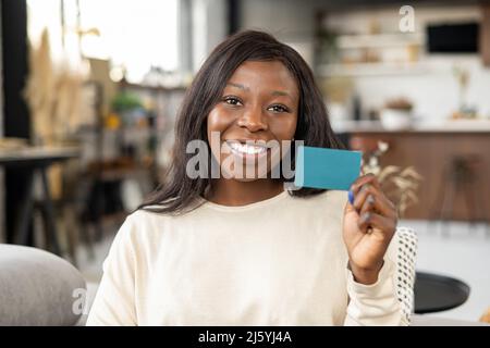 Achats en ligne. Bonne jeune femme souriant avec plaisir, assis au canapé avec une carte de crédit et regardant l'appareil photo. Bonne fille biraciale montrant carte de crédit en plastique Banque D'Images