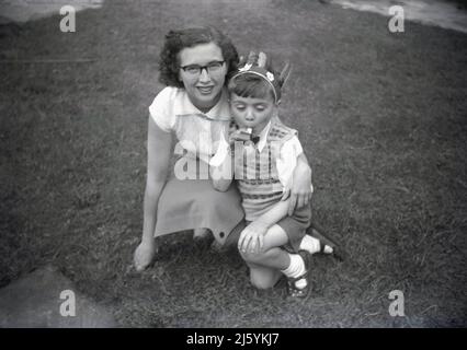 1950s, historique, à l'extérieur sur une pelouse, une mère s'agenouillant avec son jeune fils pour une photo, Angleterre, Royaume-Uni. Le petit garçon a un chapeau indien rouge et souffle un jouet ruban de fête souffleur ou sifflet, après avoir été à une fête d'enfants comme il est habillé intelligemment dans une chemise et noeud papillon et un pull sans manches ou débardeur sur le dessus, avec des shorts et des sandales. Banque D'Images