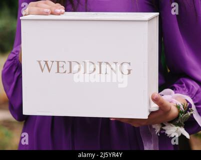 Boîte blanche avec signe Wedding. La demoiselle d'honneur en robe violette tient une boîte pour les cadeaux. Cérémonie de mariage au lilas en plein air. Fermeture de la boîte Invitatiaon Banque D'Images