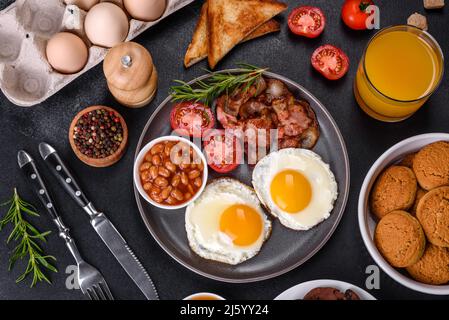 Un délicieux petit déjeuner sain et nutritif avec des œufs frits, du bacon, des haricots, un verre de jus, des biscuits d'avoine, du lait et de la confiture. Une alimentation saine au début o Banque D'Images