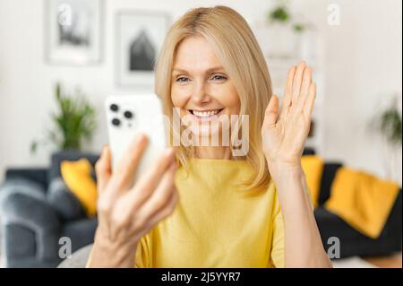 Heureuse femme mûre souriante assise dans la cuisine, se détendant et parlant à son ami ou sa famille via la connexion vidéo, femme senior tenant un téléphone mobile et en agitant la main Banque D'Images