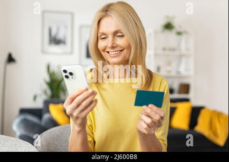 Femme mûre souriante satisfaite assise sur le canapé, tenant une carte de crédit et un smartphone, payant sur Internet, faisant l'ordre, charmante dame aime faire du shopping à la maison Banque D'Images