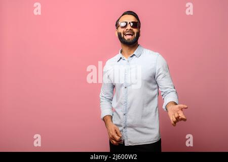 Joyeux Indien en lunettes de soleil prtend guitariste joueur, excité hispanique gars dans décontracté Jean chemise aime jouer sur la guitare imaginaire, a un plaisir, isolé sur rose Banque D'Images